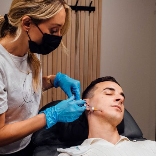 Jaw fillers. Cropped image of young man doing cosmetological injections. Male model against grey studio background. Concept of men's health, skin care, hygiene and male cosmetological treatment by Breanna Injector | Ella Esthetics in Alexandria, VA
