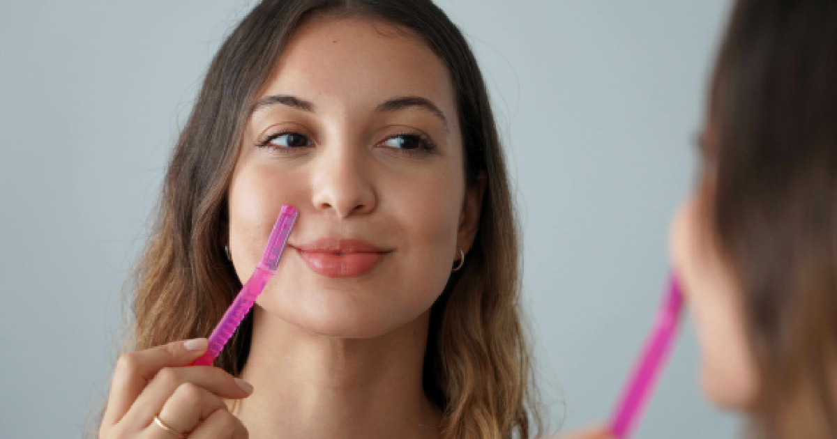 A young woman is shown performing a Dermaplaning Facial on herself with a pink razor while looking in a mirror in Alexandria, VA.